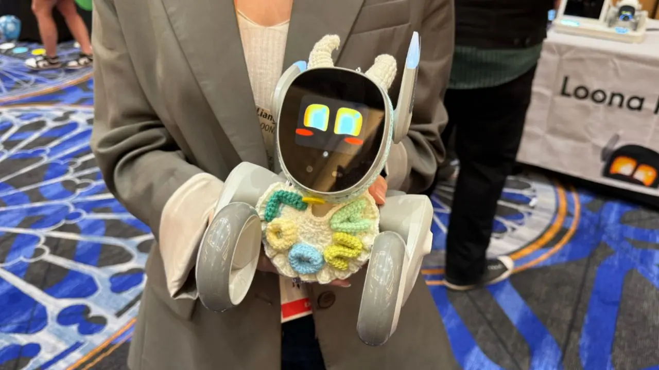 A person holds a small, cute robot with expressive digital eyes on its screen face and two wheels. The robot is adorned with a colorful knit accessory.
