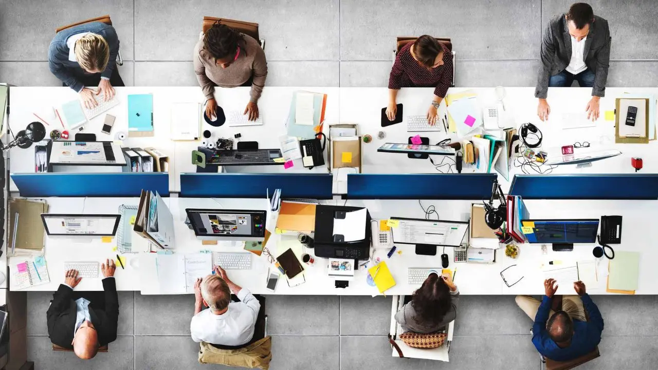 Top view of a busy office with diverse employees working at shared desks, using laptops and monitors. The scene conveys collaboration and productivity.