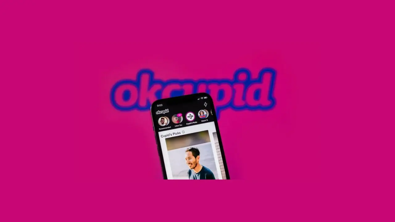 A smartphone with an OkCupid dating profile screen is shown against a vibrant pink background. The app's logo is prominently displayed, conveying a modern and social tone.