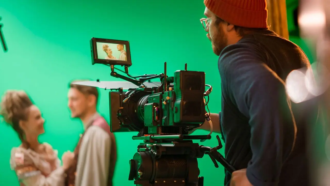 Filmmaker operates a camera on a set with a green screen background. Two blurred actors in period costumes stand in the distance, conveying a cinematic atmosphere.