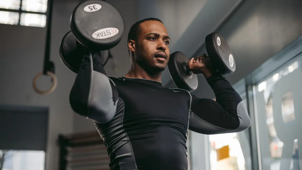 Man in a gym lifting two dumbbells at shoulder height. He wears a black athletic outfit, and his expression is focused and determined.