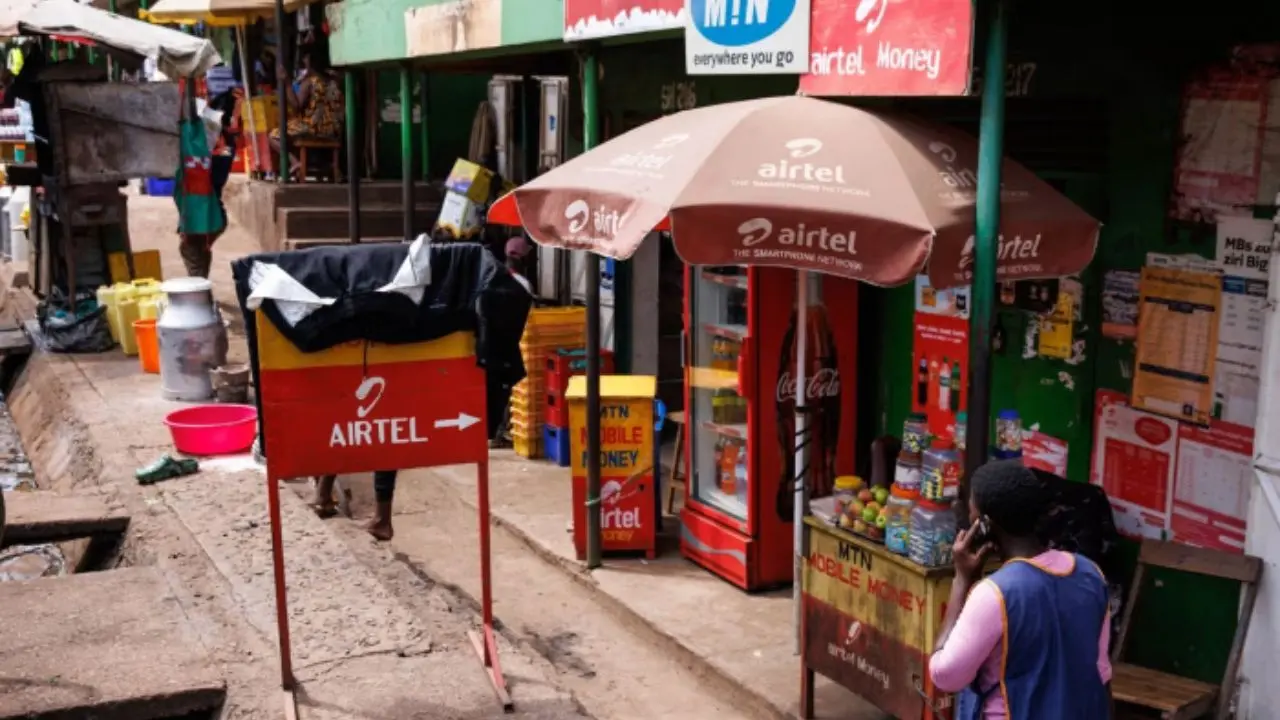A busy street stall offers Airtel services, with umbrellas, signs, and a stocked drinks fridge. A person is making a call, creating a lively atmosphere.