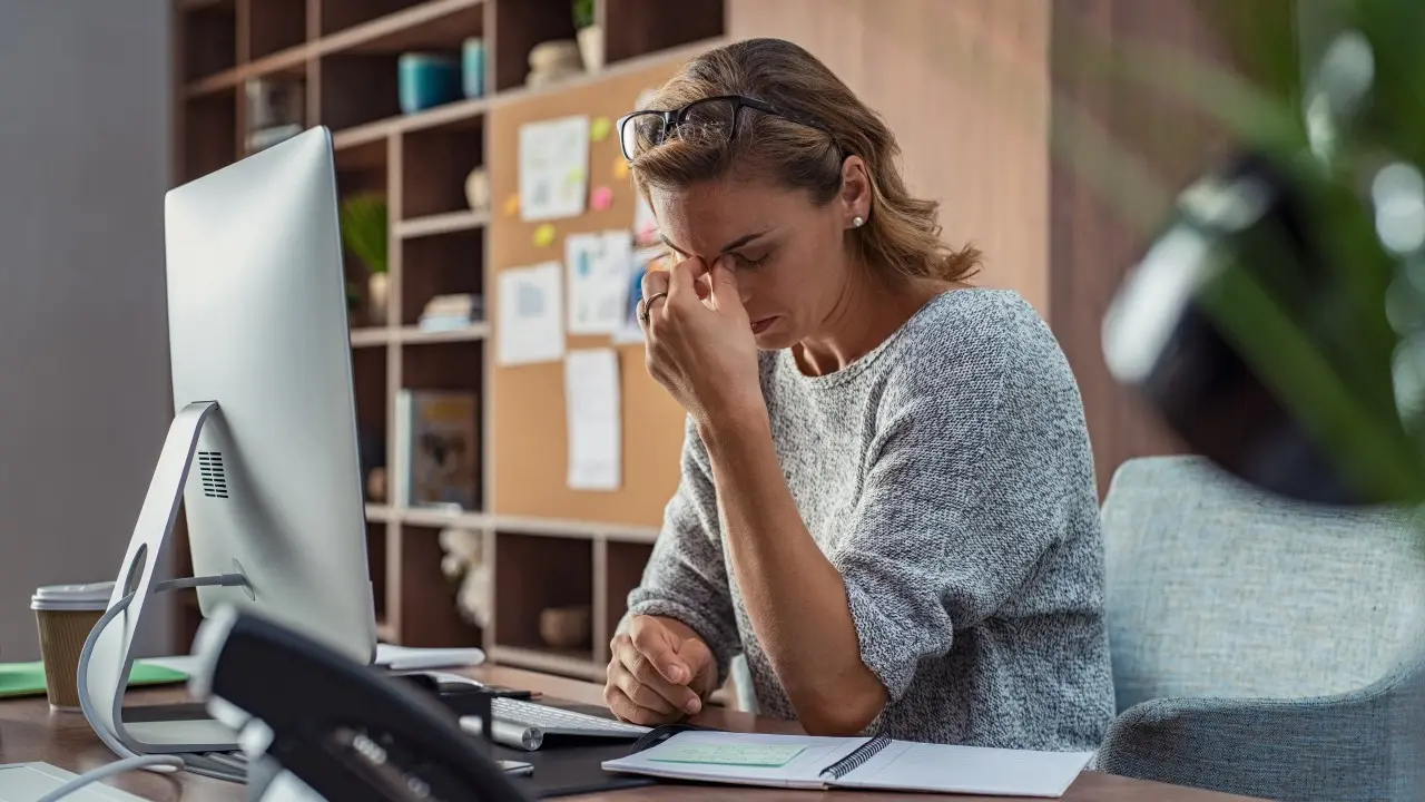 A woman in an office rubs her eyes in frustration, sitting at a desk with a computer, papers, and a shelf with files in the background.