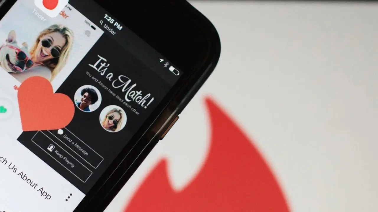 A phone displaying a Tinder "It's a Match!" screen with profile photos. A large red heart graphic and Tinder's flame logo are visible, conveying excitement.
