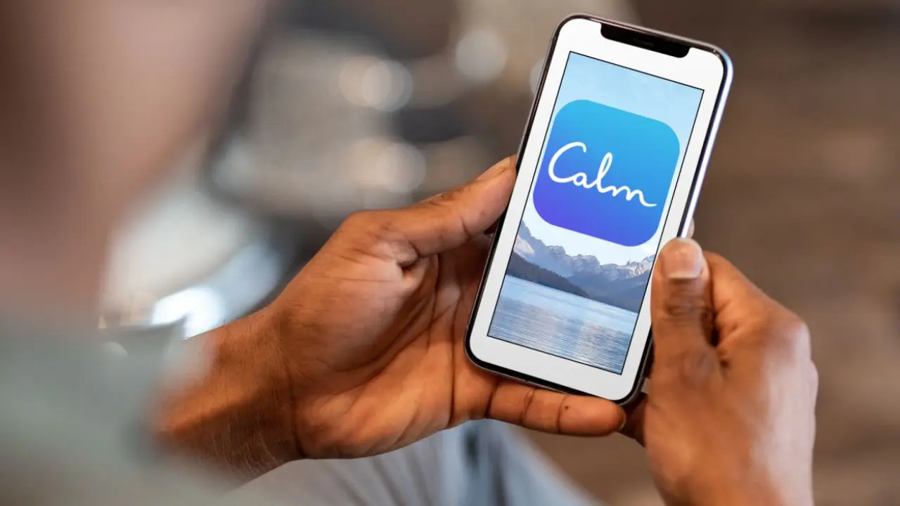 A person holds a smartphone displaying the Calm app logo against a serene mountain lake background, conveying relaxation and mindfulness.