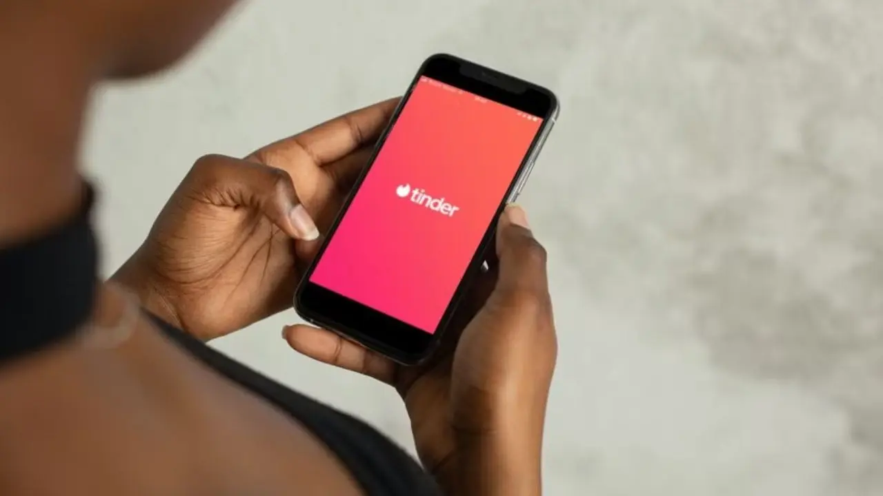 A person holds a smartphone displaying the Tinder app on the screen, set against a neutral background. The scene conveys a sense of anticipation.