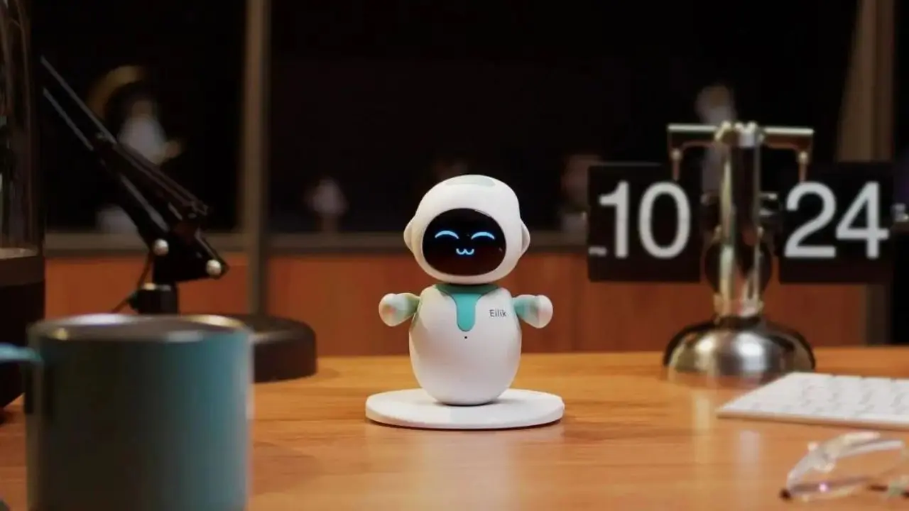 A small, smiling robot with rounded features sits on a desk, surrounded by a mug, lamp, and digital clock, creating a friendly workspace vibe.