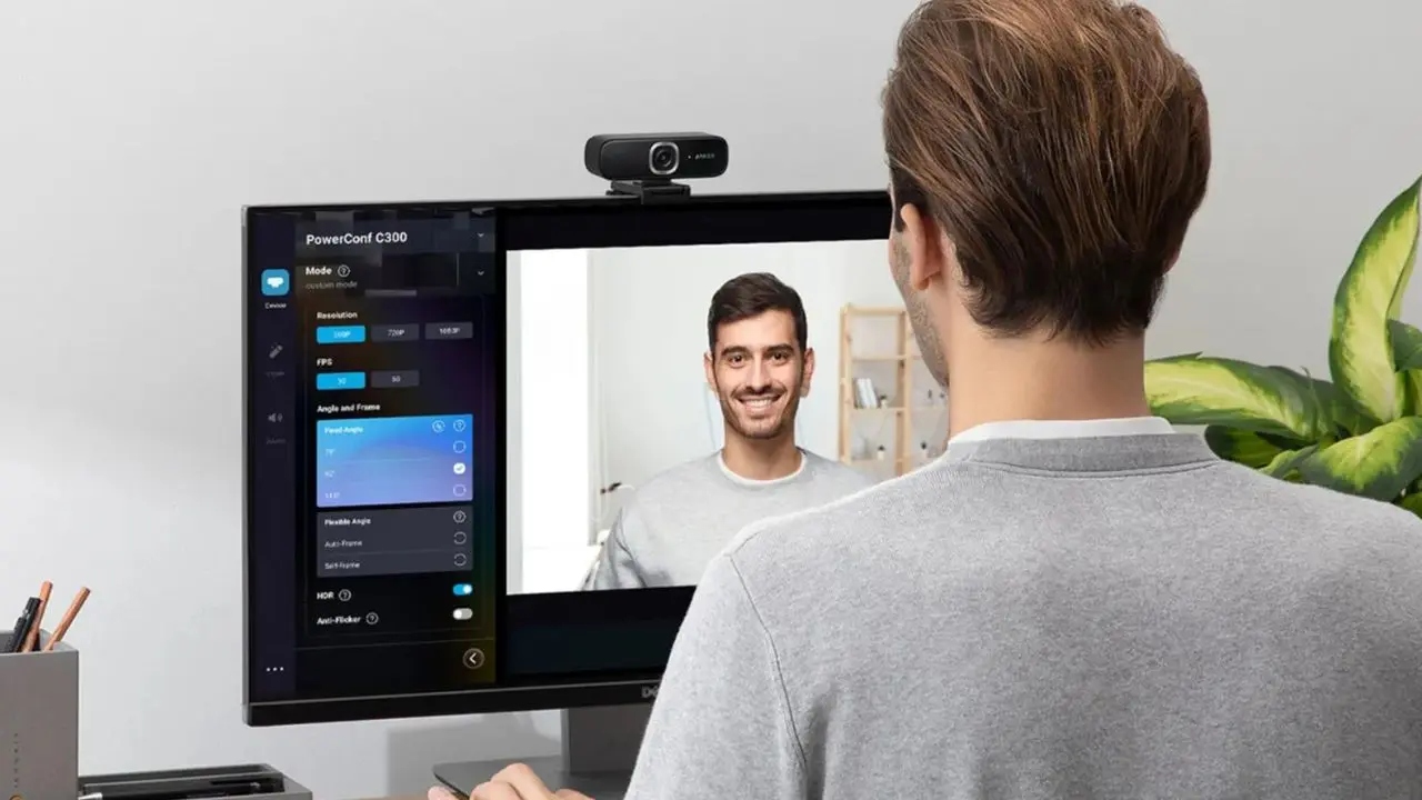 A man is sitting at a desk, facing a computer screen displaying webcam software settings on the left and his smiling image on the right. A webcam is mounted atop the monitor. The atmosphere is focused and professional, with a plant in the background.