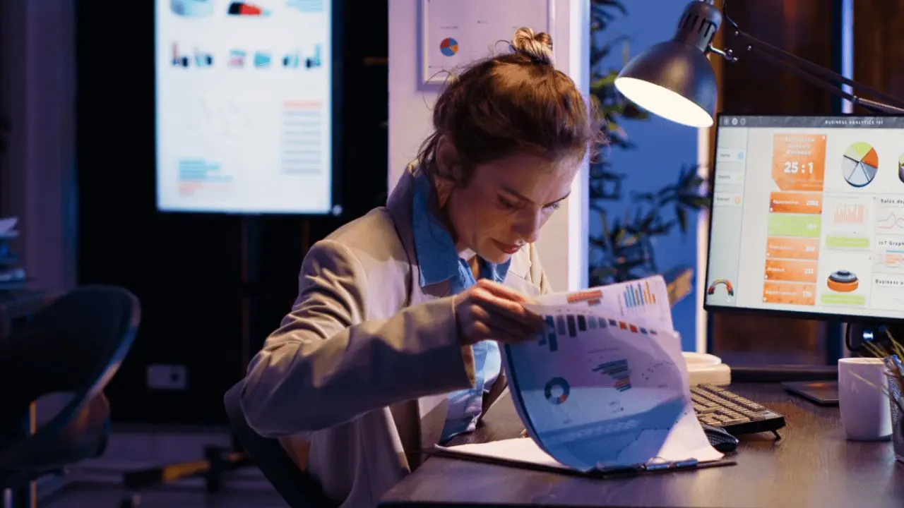 A focused woman in an office reviews printed charts at her desk, surrounded by computers displaying colorful graphs. The room is dimly lit, suggesting a late night work session.