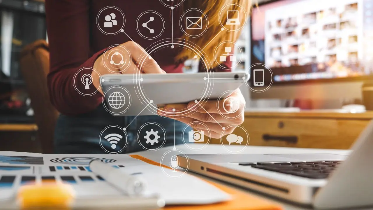 A person uses a tablet at a desk with a laptop, surrounded by digital communication icons. The setting is a modern office, evoking connectivity and technology.