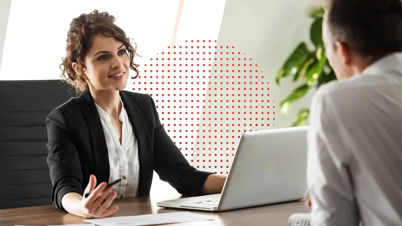 A woman in business attire talks to a man in an office. She smiles warmly, holding a pen. A laptop and documents are on the desk. A red dotted circle accents the image.