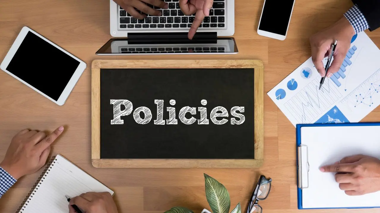 Overhead view of a workspace with a chalkboard reading "Policies" surrounded by hands working on a laptop, documents, and digital devices.
