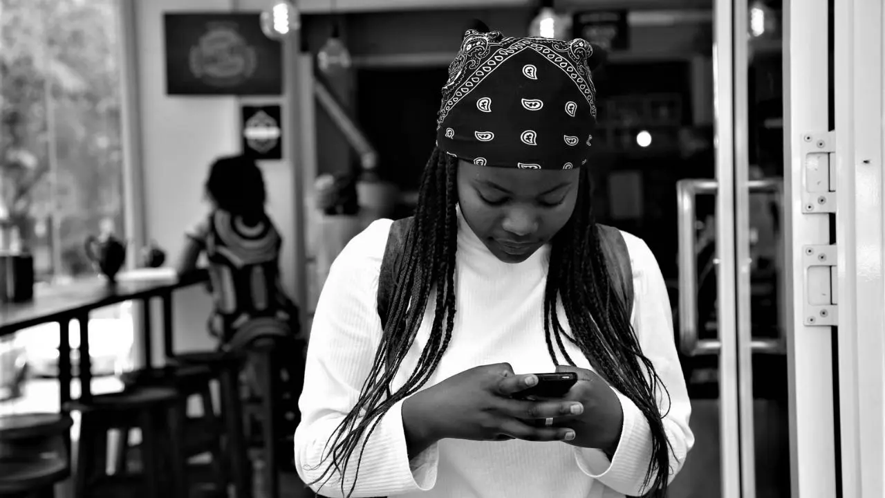 Young woman in a bandana and sweater, focused on her phone in a café. Black and white image, relaxed atmosphere with stools and blurred background.