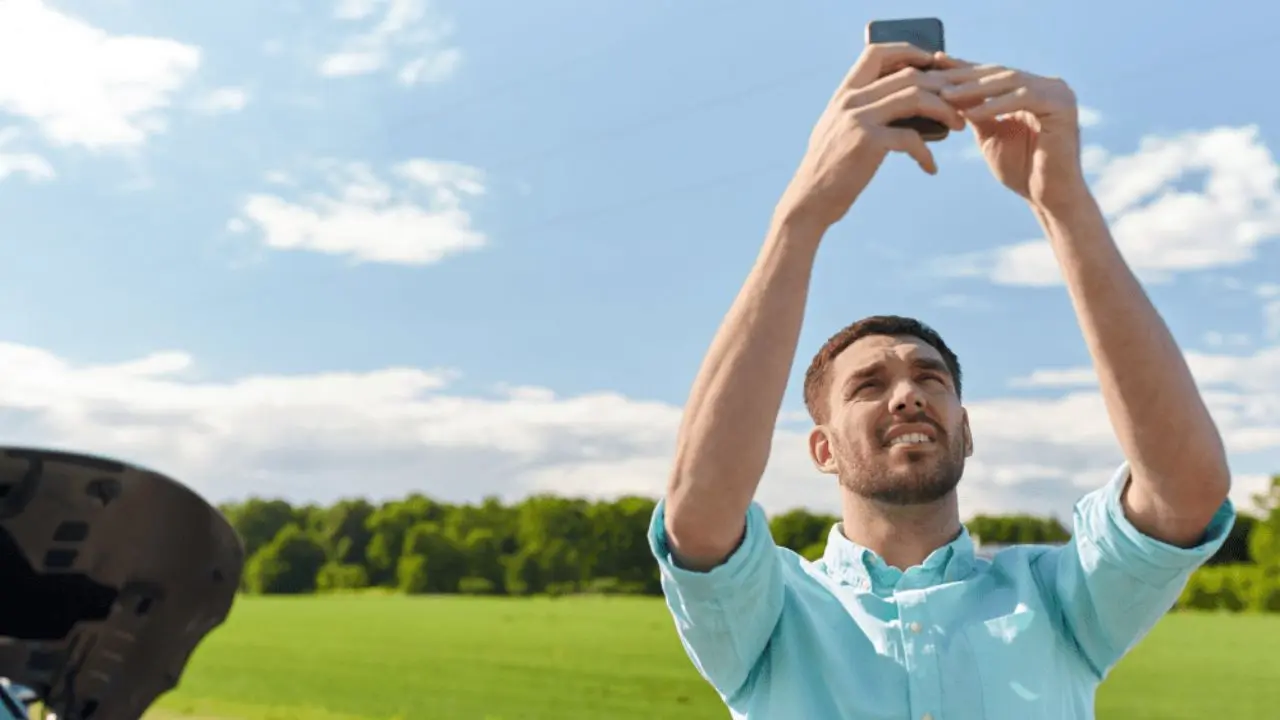 A man stands in a field with a broken-down car, holding his phone up for reception. He looks concerned, under a bright blue sky with soft clouds.