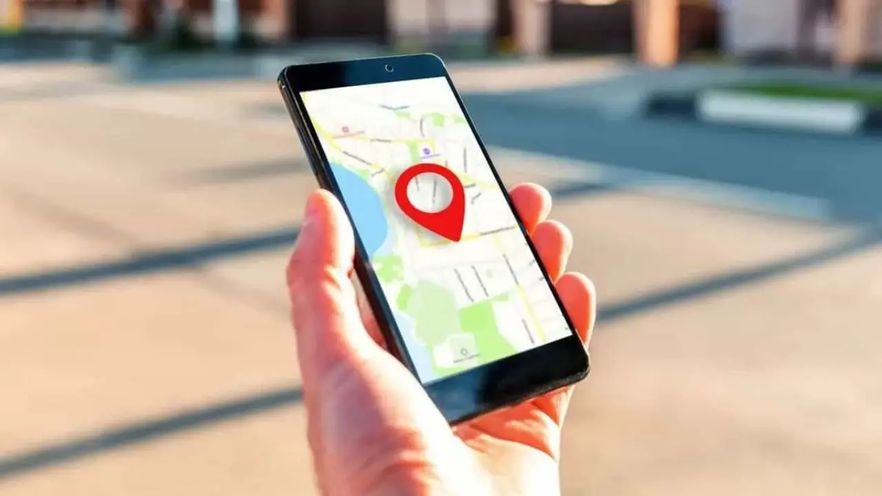 A hand holds a smartphone displaying a digital map with a prominent red location pin. The blurred outdoor background suggests a street scene.