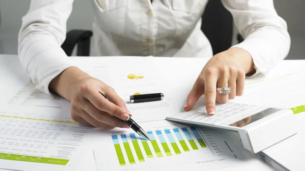 Person working with financial documents, using a calculator and pointing at a colorful bar chart. The scene conveys focus and professionalism.