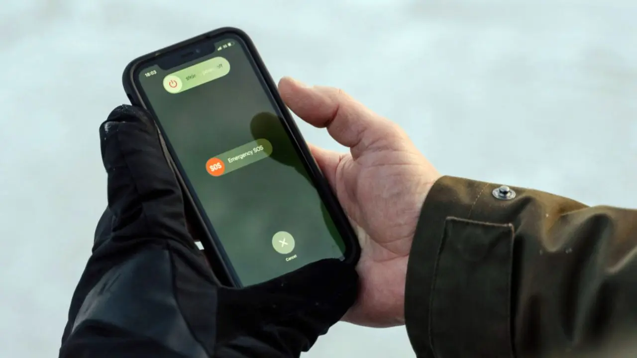 A person outdoors wearing gloves holds a smartphone displaying the Emergency SOS screen. The background is blurred, suggesting a cold environment.