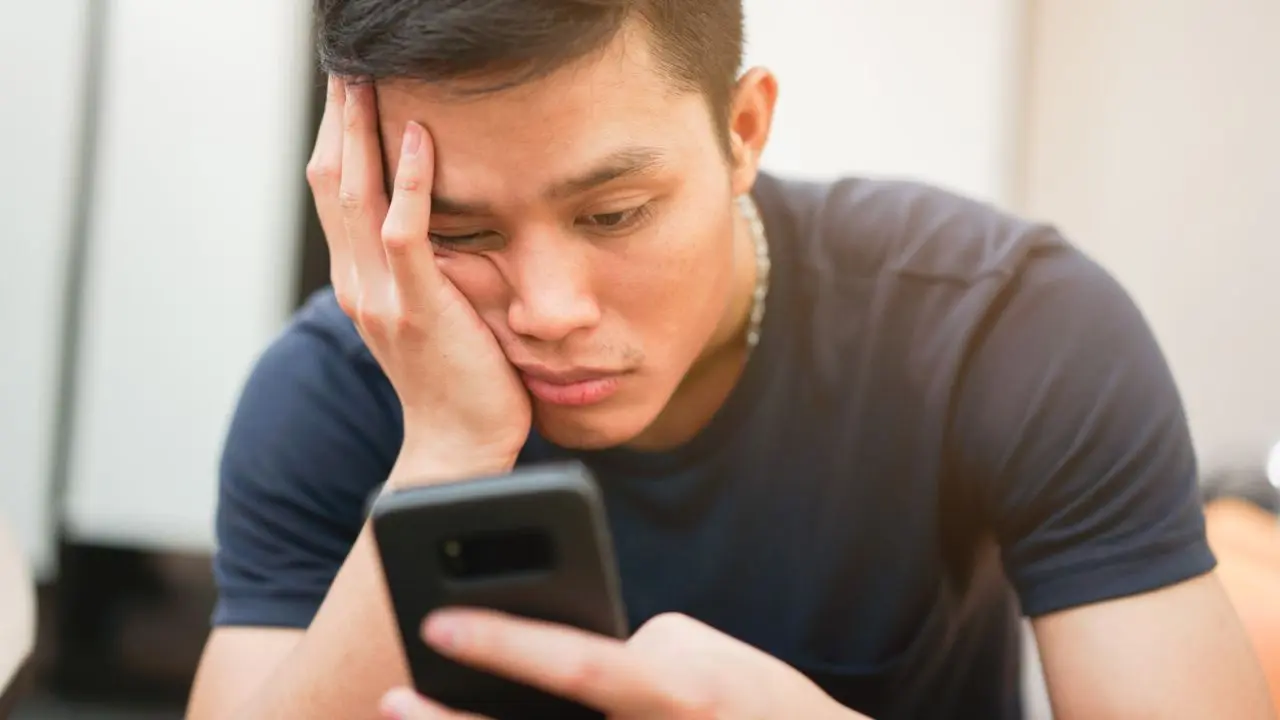 A man in a navy shirt looks at a smartphone with a bored expression, resting his head on his hand. Soft lighting creates a neutral, casual mood.