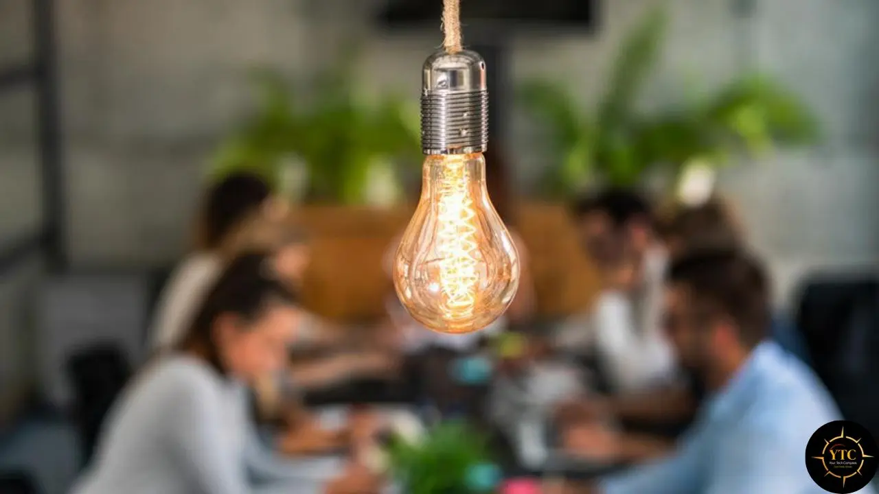 A lit Edison bulb hangs in focus above a blurred group of people in a meeting.