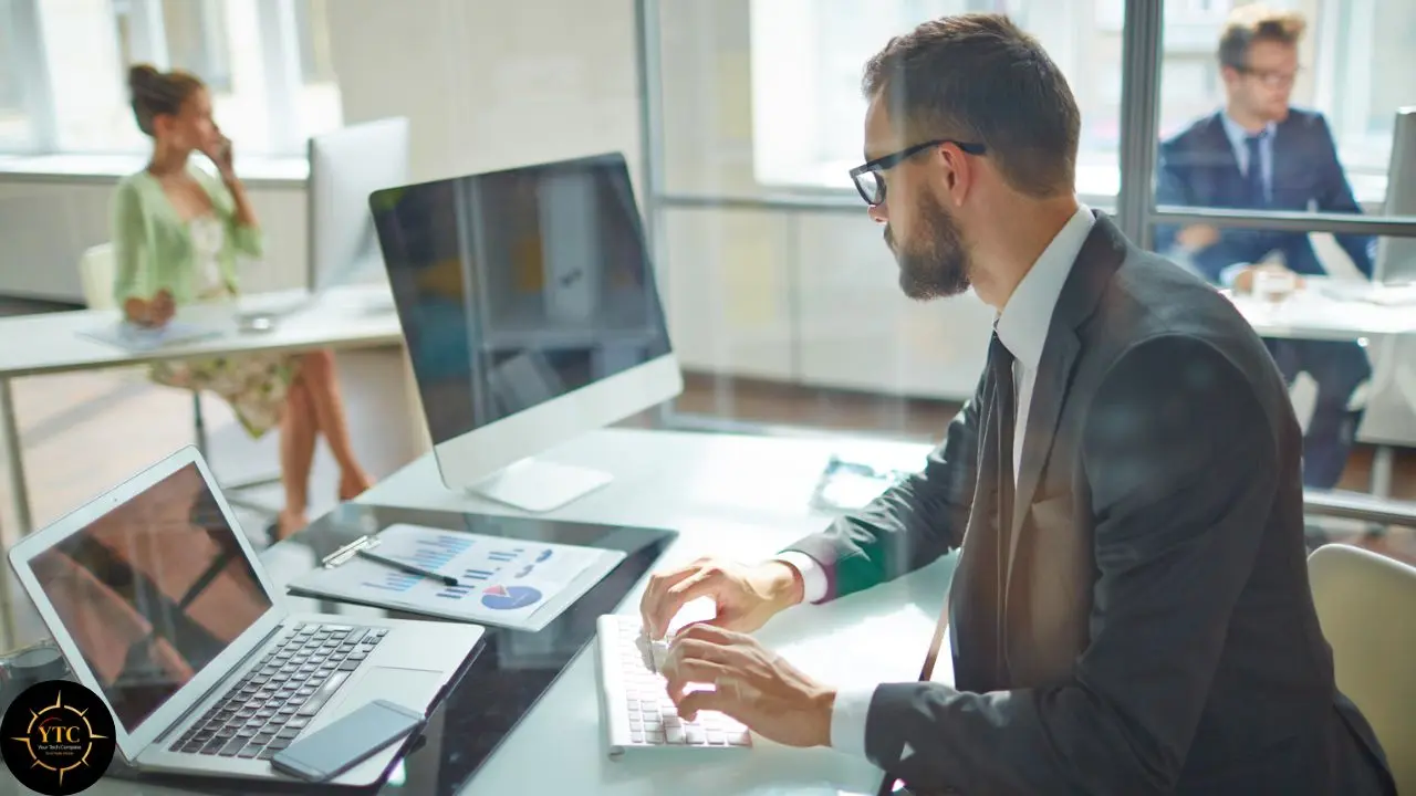 A man in a suit and glasses types on a keyboard in a modern office, with a laptop, desktop monitor, and charts on his desk.