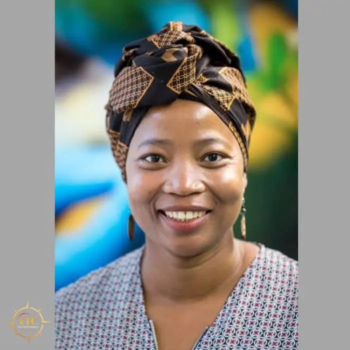 Portrait of a smiling Black woman wearing a patterned headwrap and a patterned shirt, with a colorful, blurred background.