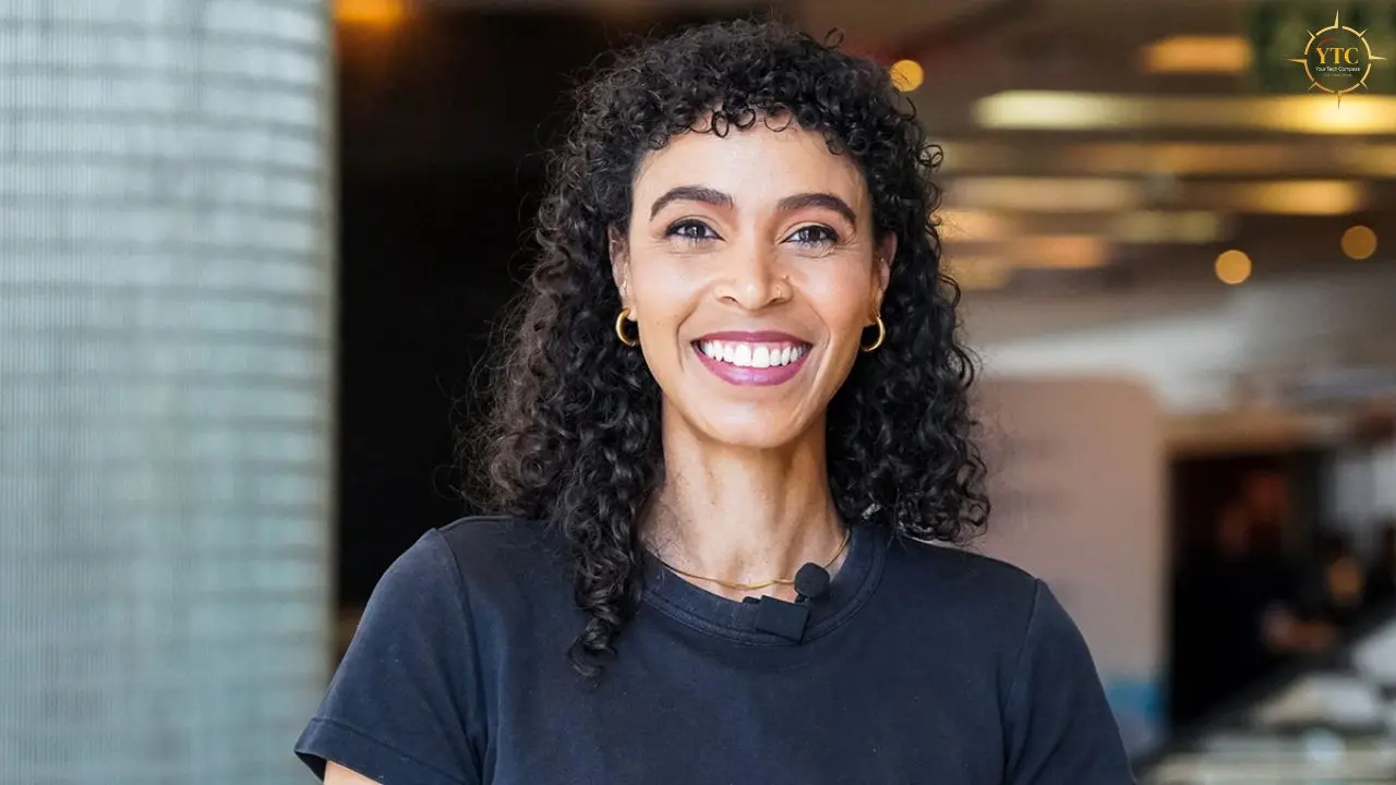 A smiling woman with curly dark hair wears a dark t-shirt and gold hoop earrings.