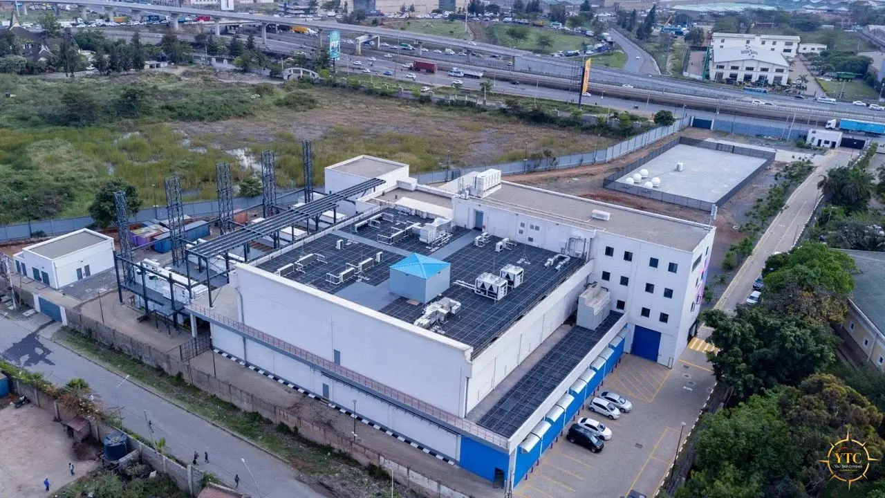 Aerial view of a large white industrial building with rooftop equipment, parking spaces, roads, and a highway in the background.