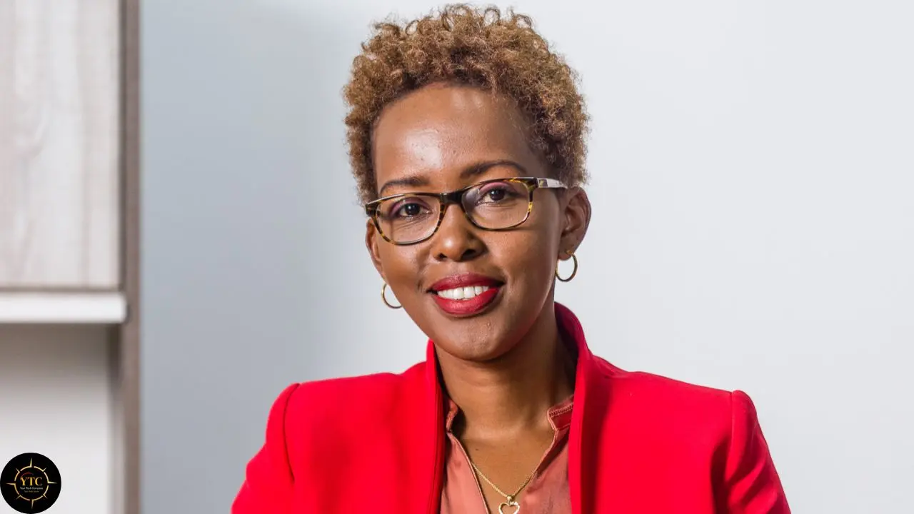 A smiling woman with short curly hair and glasses wears a bright red blazer and hoop earrings against a pale background.